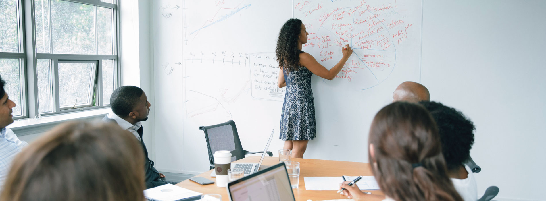 Conference room business setting with individual writing on whiteboard and peers seated.